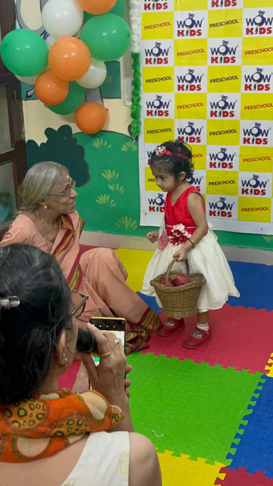 A toddler in a red party dress carrying a basket at a school celebration while her grandmother cheers her on