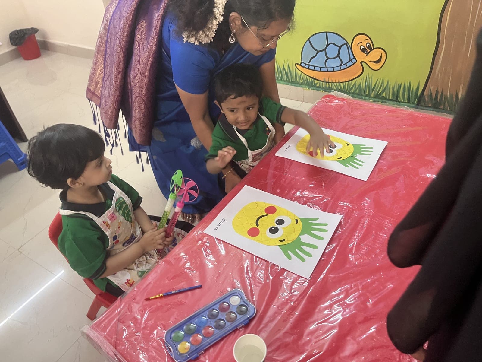 Teacher guiding two preschoolers as they paint handprint pineapple art at Wow Kids Bubbles preschool, Royapettah